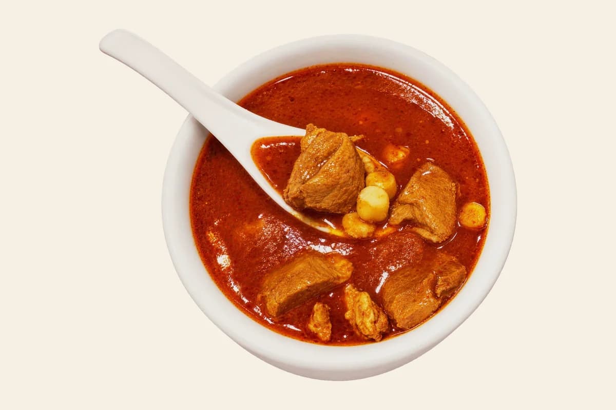 A bowl of red pozole with pork and hominy and a white spoon
